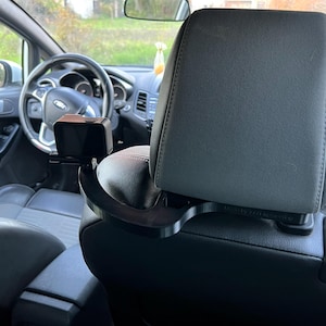 Könnte beinhalten: Schwarze Kfz-Kopfstützenhalterung mit einem rechteckigen Gerät. Die Halterung ist gebogen und so konzipiert, dass sie um die Kopfstütze passt. Der Innenraum eines Autos ist im Hintergrund sichtbar.