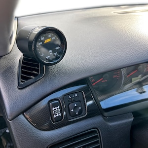 May include: Black oil pressure gauge mounted on a car dashboard. The gauge has a black face with white markings and a needle indicating the oil pressure. The dashboard is black with various buttons and vents.