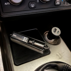 Puede incluir: Un accesorio negro para automóvil con una pantalla digital y el texto "AUTO TIMER FORNA & TURBO" se coloca sobre una alfombrilla negra. Un cargador USB está enchufado a una toma de corriente del automóvil. Se ven el salpicadero y el volante del coche.