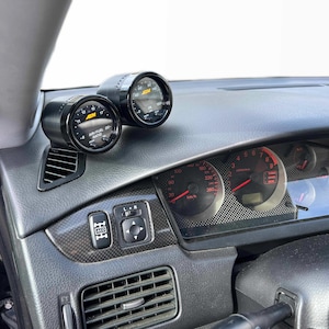 May include: Dashboard of a car with two black gauges mounted on the left side. The gauges have white and yellow markings. The dashboard also features a speedometer and other controls.