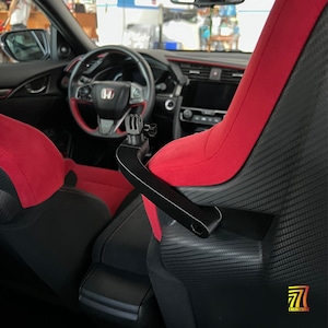 Può includere: Un supporto per fotocamera nero fissato al lato di un sedile auto rosso e nero. Il supporto presenta un'interfaccia standard a tre punte per action cam. L'interno dell'auto include un volante con logo Honda e finiture del sedile in carbonio.