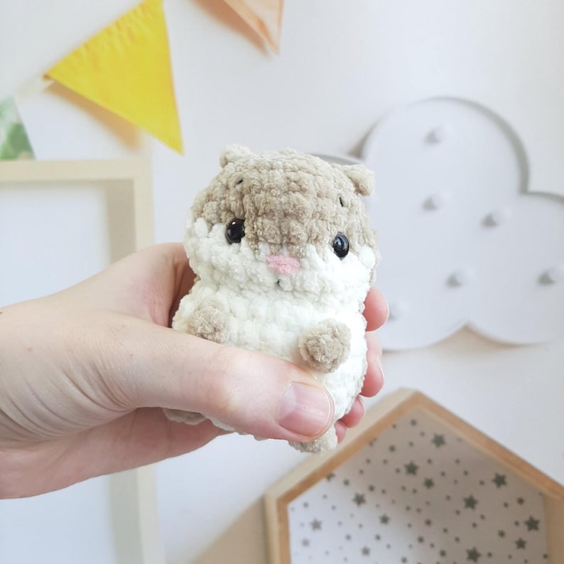 May include: A handmade crochet hamster toy in shades of beige and white, with black eyes and a pink nose. The plush toy is held in a hand, showcasing its soft, textured appearance. The hamster is a cute, cuddly item.