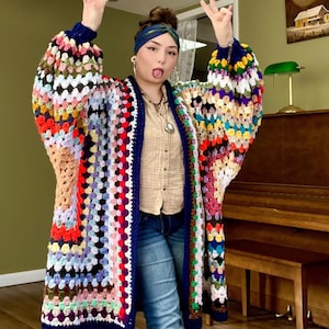 May include: A colorful, handmade crochet cardigan with a granny square design. The long, open-front sweater features a mix of vibrant colors, including red, blue, yellow, and green, with a dark blue trim. The sleeves are long and voluminous.