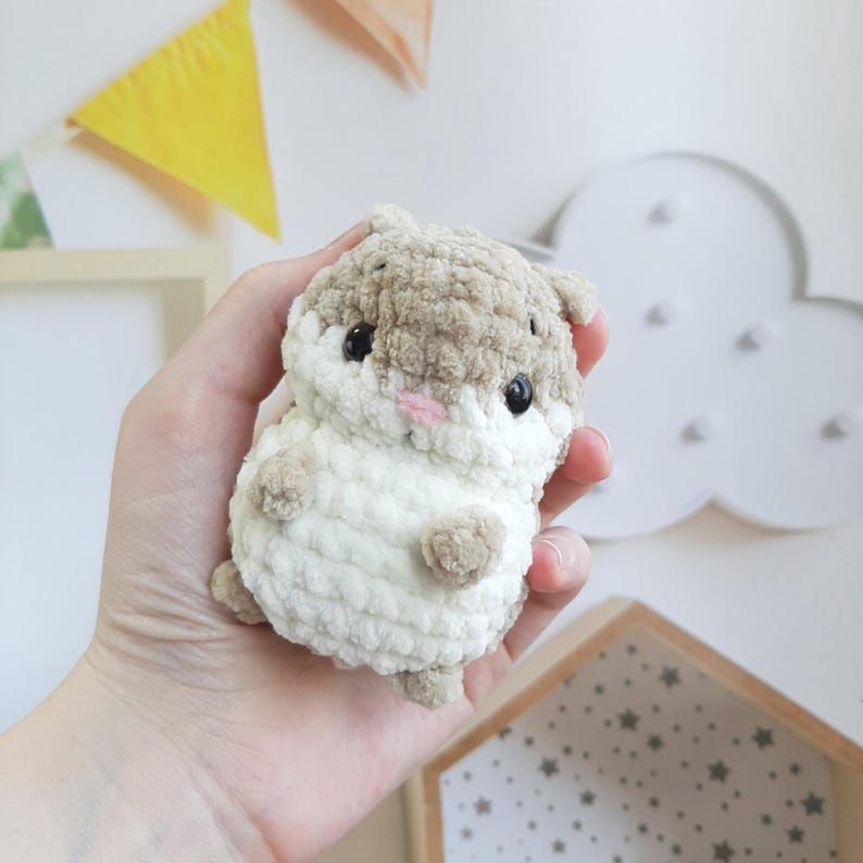 May include: A handmade, plush hamster toy in shades of brown and white. The crochet hamster has black eyes and a pink nose. The toy is held in a hand, showcasing its soft, textured appearance. A charming, cuddly stuffed animal.