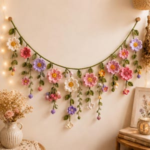 May include: A decorative wall hanging featuring a long green crocheted vine draped in a curve. Attached to the vine are various crocheted flowers in shades of pink, white, purple, and yellow, along with green leaves and small dangling flower buds.
