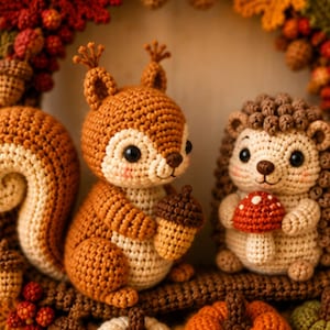 May include: A crochet squirrel holding a brown acorn and a crochet hedgehog holding a red and white mushroom, both featuring cream and brown yarn textures, positioned among crocheted autumn leaves, berries, and small pumpkins.