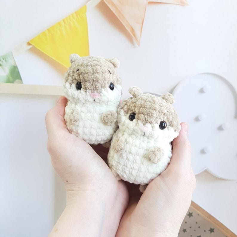 May include: Two handmade crochet hamster plushies. The plushies are beige and brown with black eyes and pink noses. The hamster toys are held in a person's hands. The background includes decorative banners.