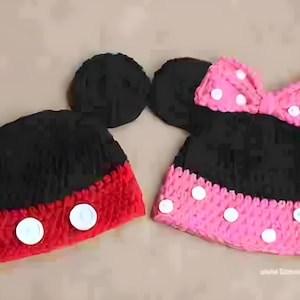 Op de afbeelding: Twee handgemaakte gehaakte mutsen, één rood en zwart met witte knopen, en de andere zwart en roze met een strik en witte stippen. Beide mutsen hebben grote zwarte oren, die lijken op cartoonkarakterhoeden.