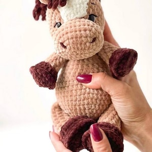 May include: A handmade crochet horse plushie in tan, brown, and white. The horse has a fluffy mane and tail, with dark brown hooves and a smiling face. The plushie is held in a person's hands.