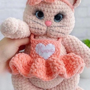 Könnte beinhalten: Handgemachte rosa Häkelkatzenpuppe mit Schleife, Stirnband und Kleid. Die Puppe hat blaue Augen mit langen Wimpern und eine rosa Nase. Das Kleid hat einen herzförmigen Aufnäher mit dem Wort "Mama". Die Puppe ist weich und plüschig.