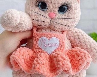 May include: A handmade pink crochet cat doll with a bow, headband, and dress. The doll has blue eyes with long eyelashes and a pink nose. The dress has a heart-shaped patch with the word "Mama" on it. The doll is soft and plush.
