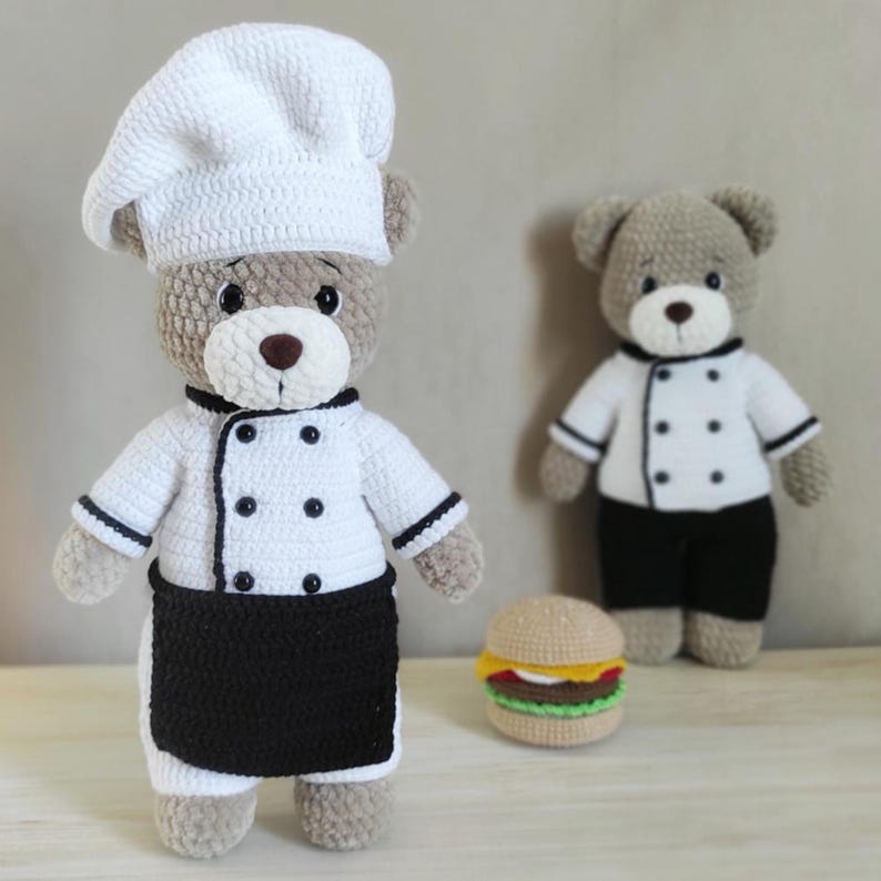 May include: Two crocheted teddy bears dressed as chefs, one with a white chef's hat, jacket, and black apron, the other with a chef's jacket and black pants. A crocheted hamburger is also present.