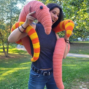 Pode incluir: Uma grande minhoca de crochê feita à mão em tons de pêssego, laranja e amarelo. A minhoca tem uma cabeça arredondada com um olho azul e seções listradas. O brinquedo de pelúcia está sendo segurado por uma pessoa em um ambiente de parque.
