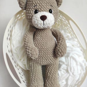May include: A handmade, plush teddy bear in a light brown color, with a white muzzle and a dark brown nose. The bear has black eyes and is standing in a white basket filled with white yarn. The bear is a soft toy.