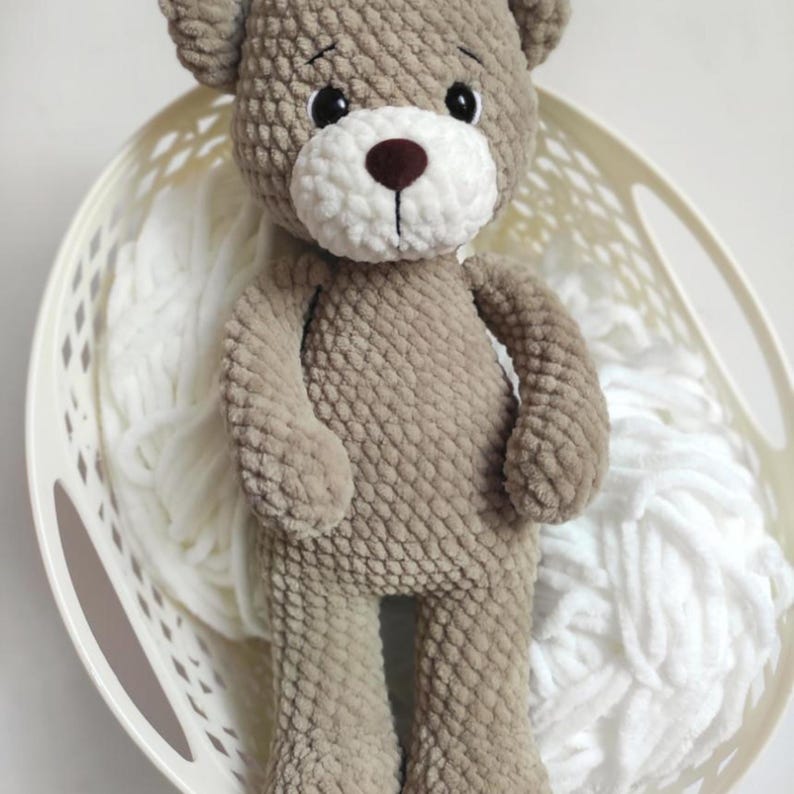 May include: A handmade, plush teddy bear in a light brown color, with a white muzzle and a dark brown nose. The bear has black eyes and is standing in a white basket filled with white yarn. The bear is a soft toy.