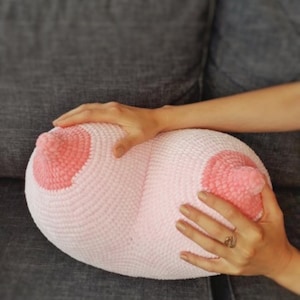 Puede incluir: Una almohada de ganchillo rosa claro con forma de senos, con pezones rosa más oscuros. La almohada está siendo sostenida por las manos de una persona, descansando sobre una superficie gris.