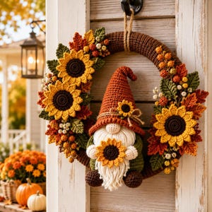 Puede incluir: Una corona de ganchillo circular con un gnomo de sombrero naranja alto, barba blanca y un girasol. La corona está decorada con girasoles, hojas verdes y bayas naranjas de ganchillo, sujeta a un aro marrón mediante un cordel de yute.