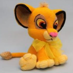 May include: A handmade crochet lion cub toy in shades of yellow, with black-lined ears and a pink nose. The lion cub has a yellow bow and is sitting down. The toy is a soft, cuddly item.