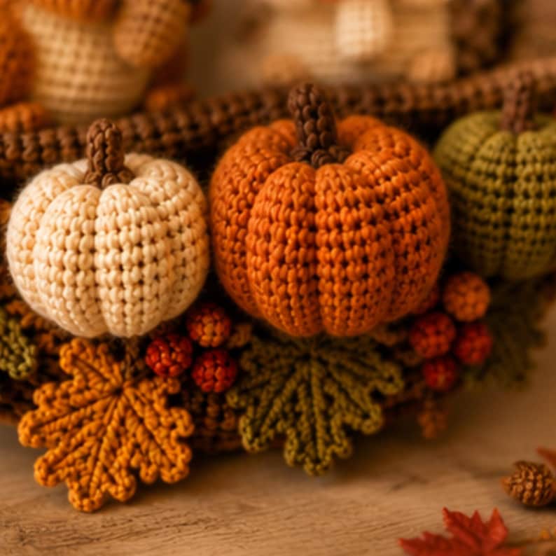 May include: A decorative wreath crafted from crocheted yarn featuring three pumpkins in cream, orange, and olive green. The arrangement includes crocheted autumn leaves in shades of gold and green, along with small red berries and brown stems.