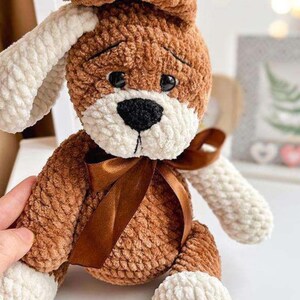 Puede incluir: Un perro de peluche hecho a mano, marr&oacute;n y blanco, con una expresi&oacute;n triste. El perro tiene una nariz negra, ojos negros y una cinta marr&oacute;n alrededor del cuello. El peluche est&aacute; hecho de hilo suave y texturizado.