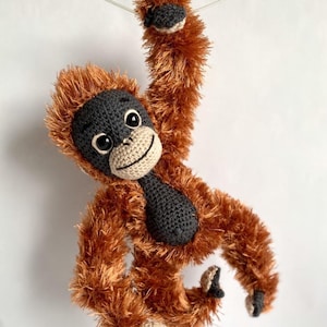 May include: A handmade crochet orangutan doll with brown fur, a black face, and a cream-colored mouth. The doll is hanging from a thin, white string. The orangutan has a friendly expression and is a soft toy.