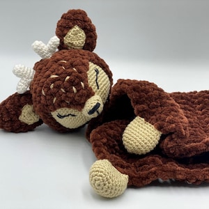 May include: A handmade brown crochet deer plushie with cream-colored accents. The deer has white antlers, a cream-colored face, and a matching blanket. The plushie is designed to be soft and cuddly.