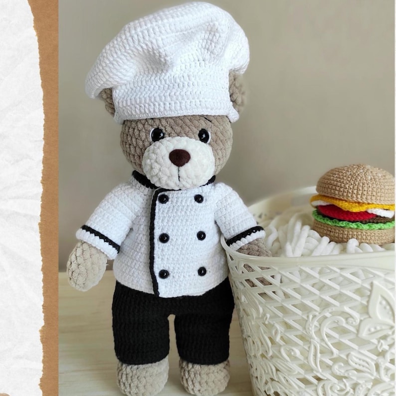 May include: A crocheted teddy bear dressed as a chef, wearing a white chef's hat and jacket with black trim and buttons, and black pants. A crocheted hamburger sits nearby in a white basket. The bear is brown and beige.
