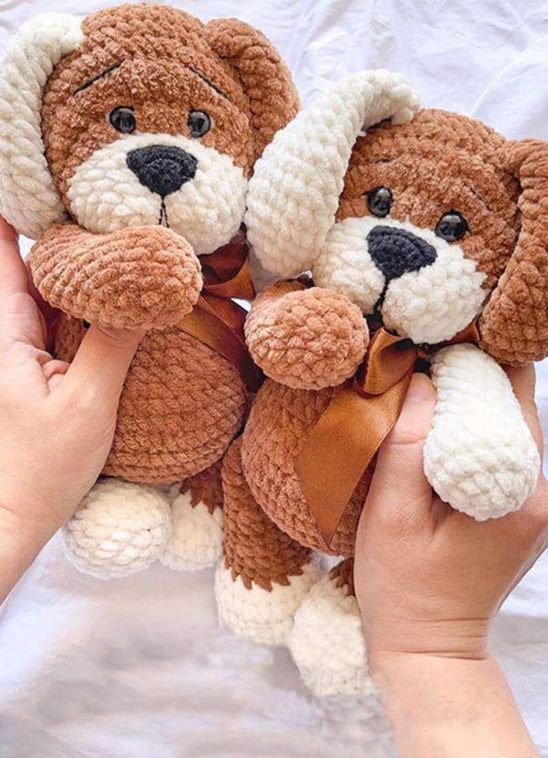 Puede incluir: Dos peluches de perro hechos a mano con ganchillo. Los perros son marrones con detalles blancos en las orejas, patas y hocico. Cada perro tiene una nariz negra, ojos oscuros y una cinta marr&oacute;n alrededor del cuello. Los juguetes son suaves y afelpados.