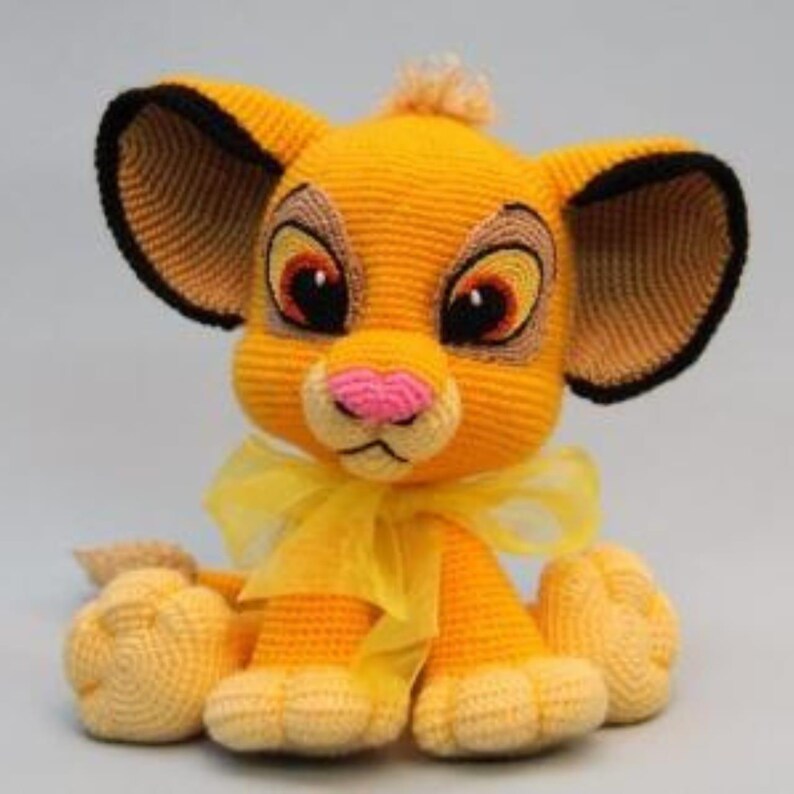 May include: A handmade crochet lion cub toy in shades of yellow, tan, and black. The lion cub has large eyes, a pink nose, and a yellow bow. The toy is made of yarn and has a soft, textured appearance.