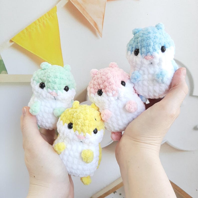 May include: Four handmade crochet hamster plushies in pastel colors: light blue, pink, yellow, and green. Each plushie has black eyes and a white belly. The plushies are held in a person's hands.