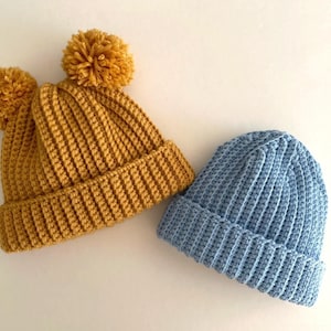 May include: Two knitted beanies. One is mustard yellow with a large pom-pom, the other is light blue. Both hats have a ribbed design. The hats are made of a soft, warm material, perfect for cold weather.