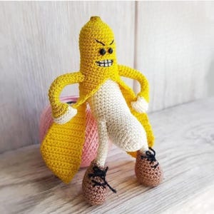 Puede incluir: Una figura de plátano de ganchillo hecha a mano con una expresión enfadada. La cáscara del plátano es amarilla y el cuerpo es blanco. Tiene zapatos marrones con cordones negros y guantes blancos. La figura está de pie en pose heroica.