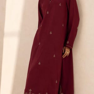 Op de afbeelding: Bordeauxrode lange tuniek en bijpassende broek met wijde pijpen. De tuniek heeft delicate gouden borduursels langs de zoom, manchetten en verspreid over de voorkant. De outfit heeft een mandarijnkraag en een zijsplit. Het model draagt gouden oorbellen en ringen.