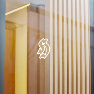 Könnte beinhalten: Ein weißer Vinyl-Aufkleber mit einem stilisierten Buchstaben S in einer gotisch inspirierten Schriftart, der auf einer transparenten Glasoberfläche angebracht ist. Das Design besteht aus einer klaren weißen Kontur auf dem Glas.