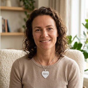 Könnte beinhalten: Eine silberne herzförmige Halskette mit dem eingravierten Wort "Mom". Die Halskette wird von einer Person getragen, die einen beigen Pullover trägt. Der Hintergrund umfasst ein Bücherregal und eine Pflanze.