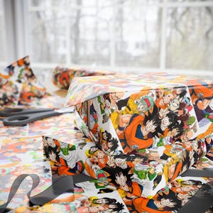Könnte beinhalten: Geschenkboxen, verpackt in buntem Geschenkpapier im Dragon Ball Z-Design. Das Papier zeigt verschiedene Charaktere in Actionposen. Schere und eine Papierrolle sind sichtbar.