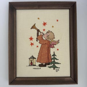 May include: A framed cross-stitch embroidery on cream fabric featuring a figure in a long orange robe playing a golden trumpet. The scene includes a small lantern, a green pine tree, and red star-shaped accents. The text 'Hummel' and 'Judy '82' is visible.