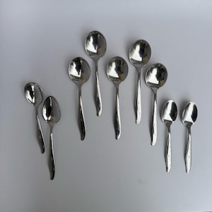 May include: A collection of nine shiny, silver spoons. The spoons have a classic design with rounded bowls and slender handles. The spoons are arranged on a white surface, reflecting the light.
