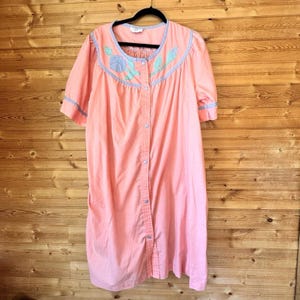 May include: A peach-colored house dress featuring short sleeves, a round neckline, and a front button-down closure. The garment is accented with light blue trim along the collar and sleeves, and includes embroidered floral details in blue and green.