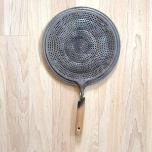 May include: A circular metal splatter screen featuring a fine mesh grid pattern across the surface. The tool includes a straight, light-colored wooden handle attached to the base, showing signs of wear and discoloration near the metal connection point.