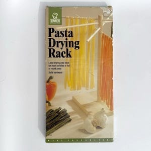 May include: A rectangular cardboard box for a Rowoco Pasta Drying Rack. The packaging features a graphic of a wooden rack holding yellow and orange pasta strands, with a red bell pepper, garlic bulbs, and green pasta noodles shown below.