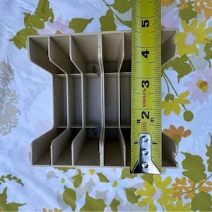 Op de afbeelding: Een beige plastic opbergorganizer met vier verticale vakken, geplaatst naast een geel Stanley-meetlint dat een hoogte van ongeveer 14 centimeter aangeeft voor de zijpaneelstructuur van de eenheid.
