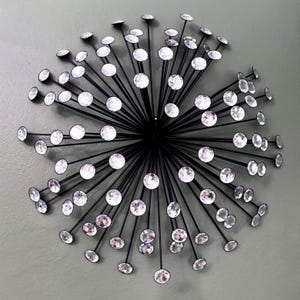 May include: A starburst wall decoration featuring numerous thin black metal rods radiating from a central point. Each rod is tipped with a clear, faceted circular crystal that reflects light, creating a spherical, sunburst-like arrangement.
