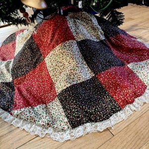 May include: A circular tree skirt featuring a patchwork design of square fabric panels in red, black, and cream colors, each decorated with a small leaf pattern. The edge of the skirt is finished with a white lace trim border.