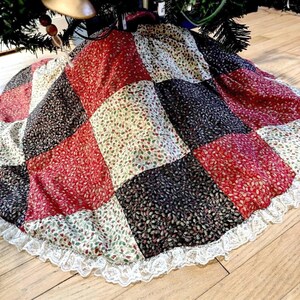 May include: A circular Christmas tree skirt featuring a patchwork design of square fabric panels in red, black, and cream colors, all decorated with a small holly leaf and berry print. The edge is finished with a white lace trim.