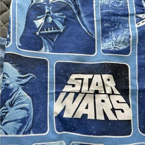 Op de afbeelding: Een blauwe stof met een herhalende Star Wars-print met witte tekst. Het ontwerp bevat grafische panelen met Darth Vader, de Death Star en Yoda tegen een donkerblauwe ruimteachtergrond met witte sterren door het hele patroon heen.