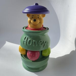 May include: A Winnie the Pooh toy featuring a yellow Pooh bear figure emerging from a green honey pot. The honey pot has the word "Honey" in green lettering. The toy includes a purple lid and colorful fruit-shaped accents.