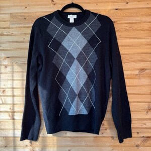 Peut inclure: Pull noir avec un motif losanges gris et blanc. Le pull a un col rond et des manches longues. Le motif losanges est centré sur le devant du pull.