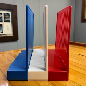 May include: A set of three bookends in the colours of the French flag: blue, white, and red. Each bookend has a rectangular base and a vertical, ridged panel. The bookends are made of plastic and are designed to hold books upright.