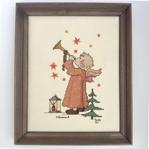 May include: A framed cross-stitch embroidery on cream fabric featuring a child in a long orange robe playing a golden trumpet. The scene includes red star accents, a small green pine tree, a lantern with a red candle, and the signature Judy '82.